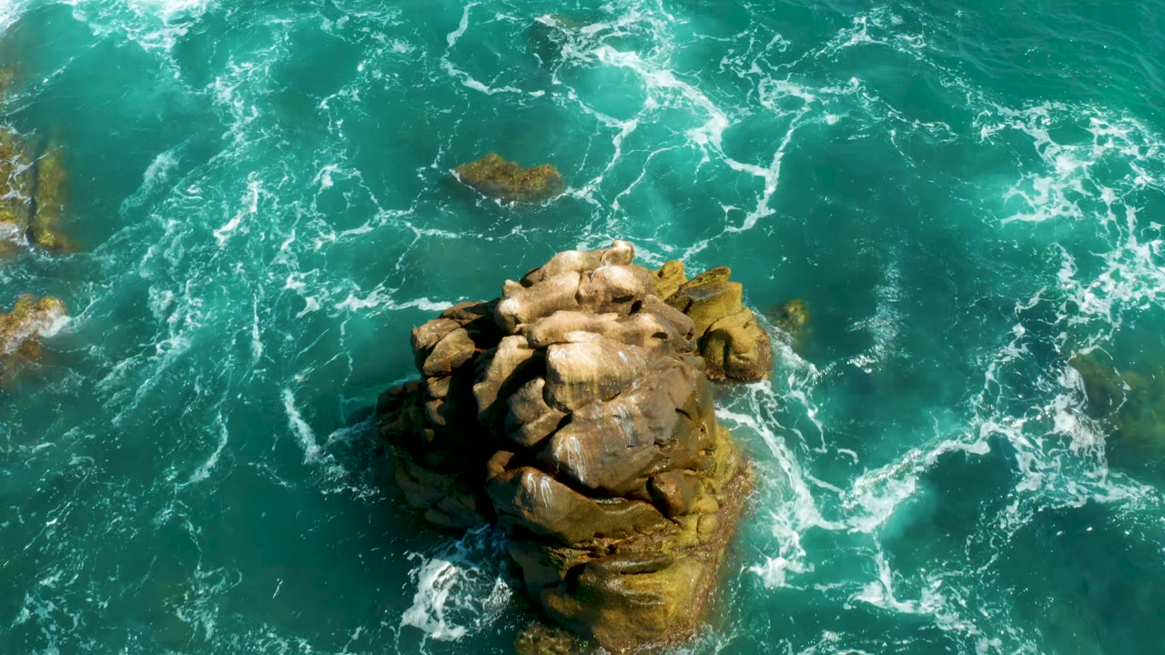 At Hilton Los Cabos, Mexico, the rock formation remains centered low in frame while white foam trails shift across the turquoise water, with no visible people.