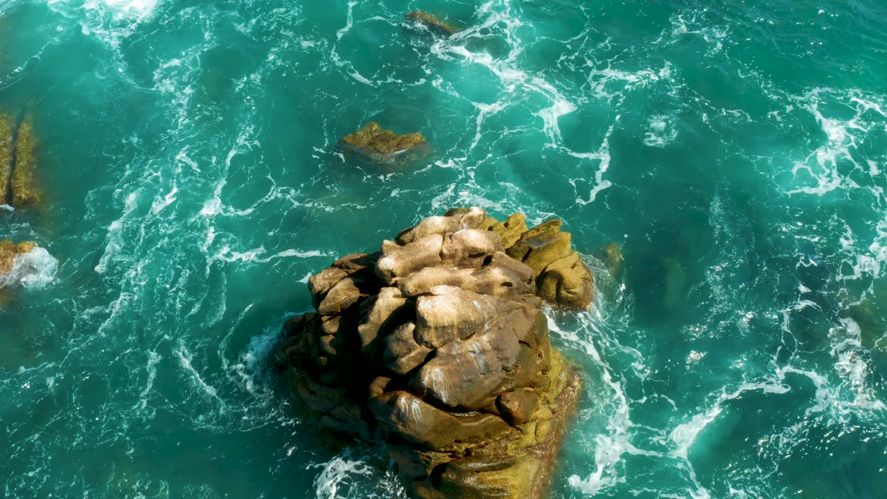At Hilton Los Cabos, Mexico, the drone view shifts so the tan rock sits farther right, with a small rock above it and whitecaps breaking through the turquoise water, with no visible people.