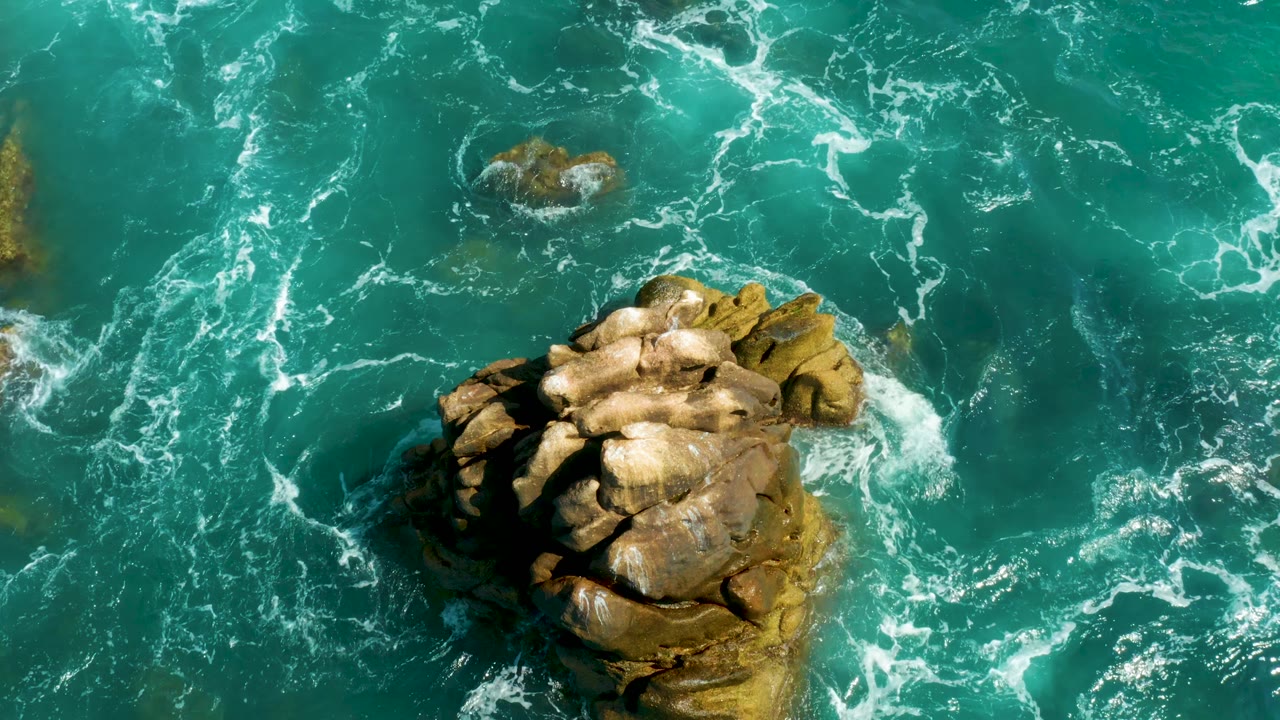 At Hilton Los Cabos, Mexico, the large tan rock sits lower left with a smaller rock above it, surrounded by turquoise water and scattered white foam, with no visible people.