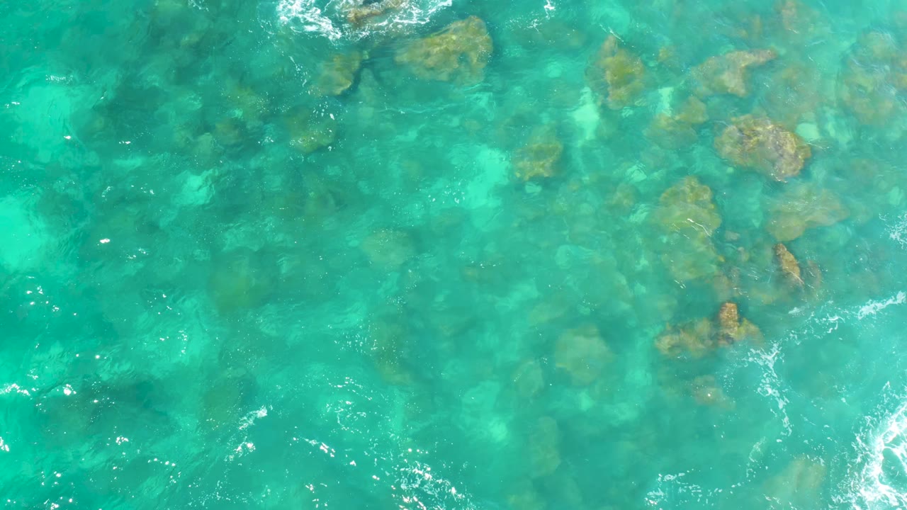 Aerial frame of clear blue-green water at Hilton Los Cabos, Mexico, with rocks visible beneath shifting ripples and no visible people.