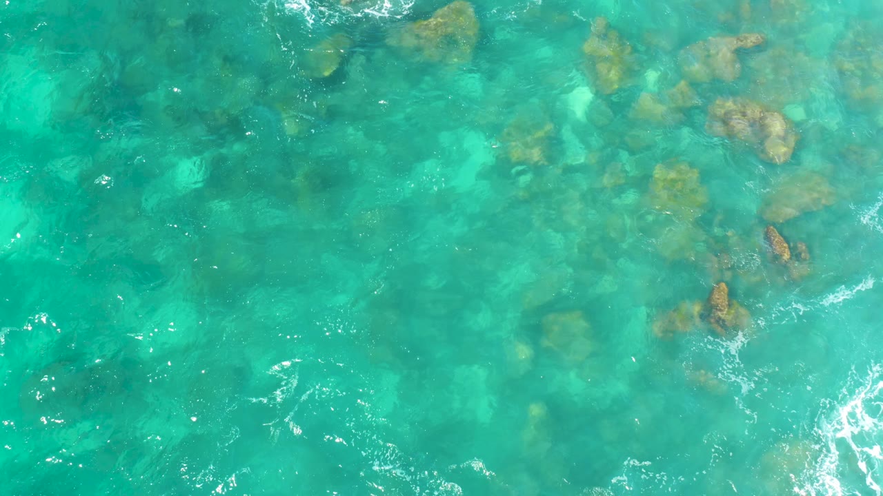 Top-down aerial view of shallow turquoise sea at Hilton Los Cabos, Mexico, with scattered underwater rocks toward the top and no visible people.