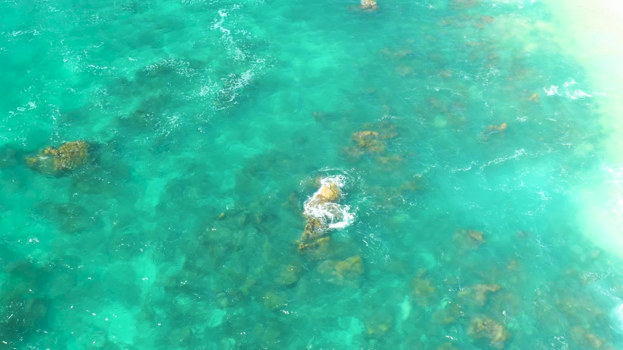 Aerial-looking frame of Hilton Los Cabos turquoise ocean, where a small foam break sits over central rocks and darker reef shapes show through the water, with no visible people.
