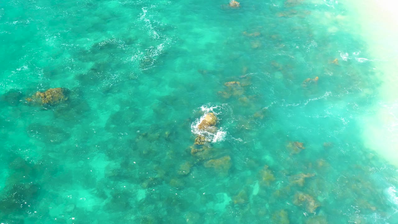 Transparent turquoise water at Hilton Los Cabos reveals scattered submerged rocks, a white foam patch near center, and a strong bright area along the right edge, with no visible people.