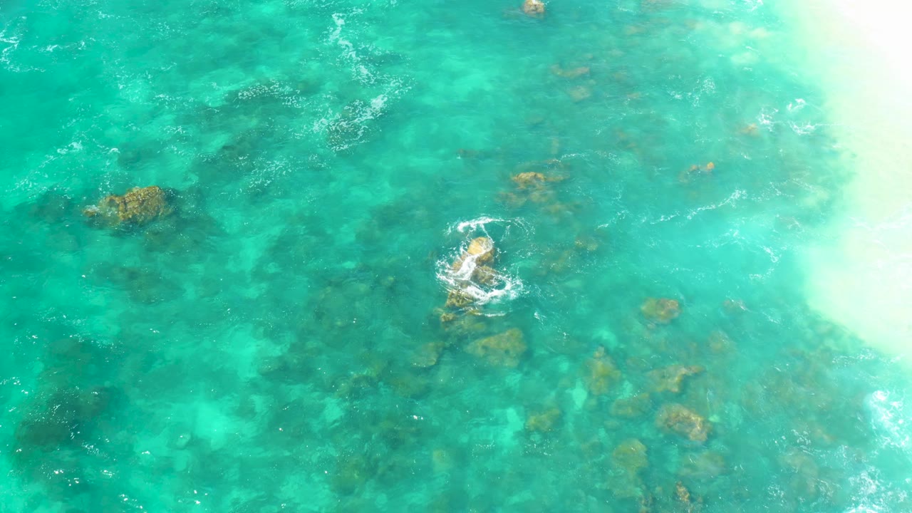 Hilton Los Cabos water appears as a top-down turquoise field, with a small circular foam pattern over rocks and pale sunlit water at right, with no visible people.