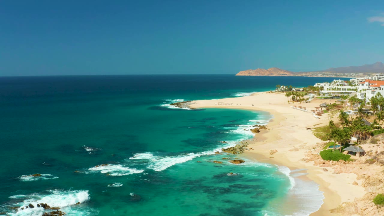 At 0:01, the Hilton Los Cabos shoreline remains in a wide aerial view as white surf spreads around offshore rocks, with beach sand, coastal buildings, distant hills, and no visible people.