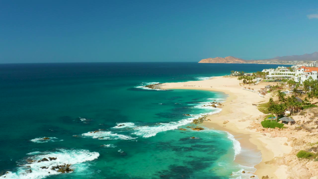 At 0:03, the composition holds a broad Hilton Los Cabos beach view with deep blue water to the left, foamy surf in the lower frame, buildings to the right, and no visible people.
