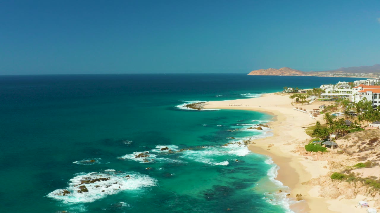 Aerial view of Hilton Los Cabos coastline with turquoise water, white surf around rocks, sandy beach, resort buildings at right, distant land on the horizon, and no visible people.