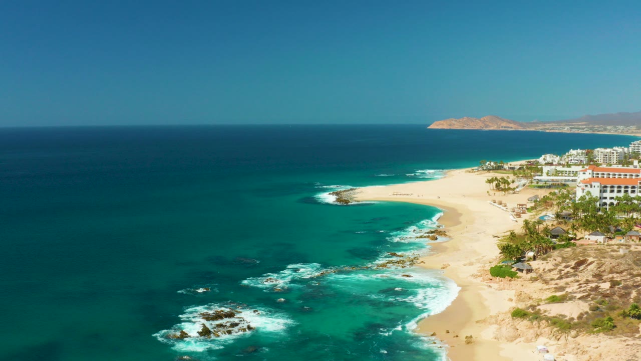 Opening frame of Hilton Los Cabos in Mexico with turquoise water at left, white waves around dark rocks, sandy beach curving past resort buildings, distant hills, and no visible people.