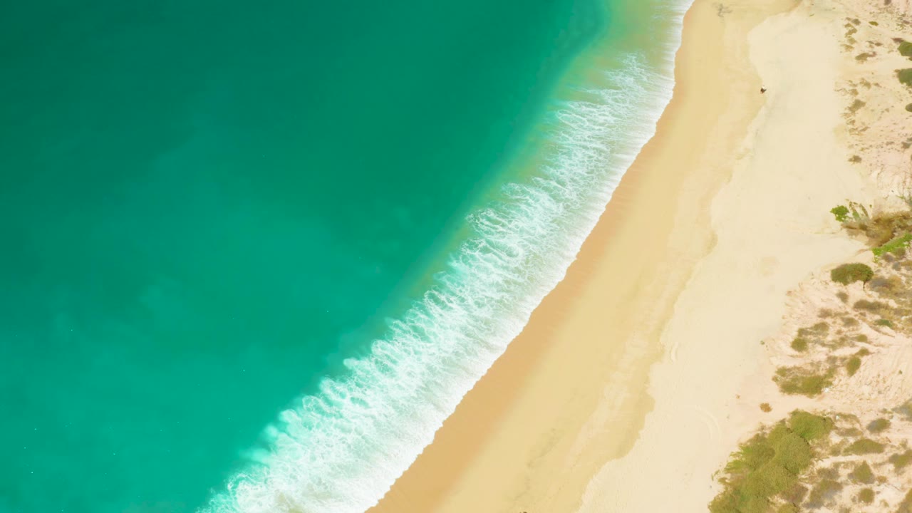Overhead Hilton Los Cabos shoreline frame with deeper turquoise water, foamy surf advancing along the diagonal beach edge, pale sand, and no visible people.