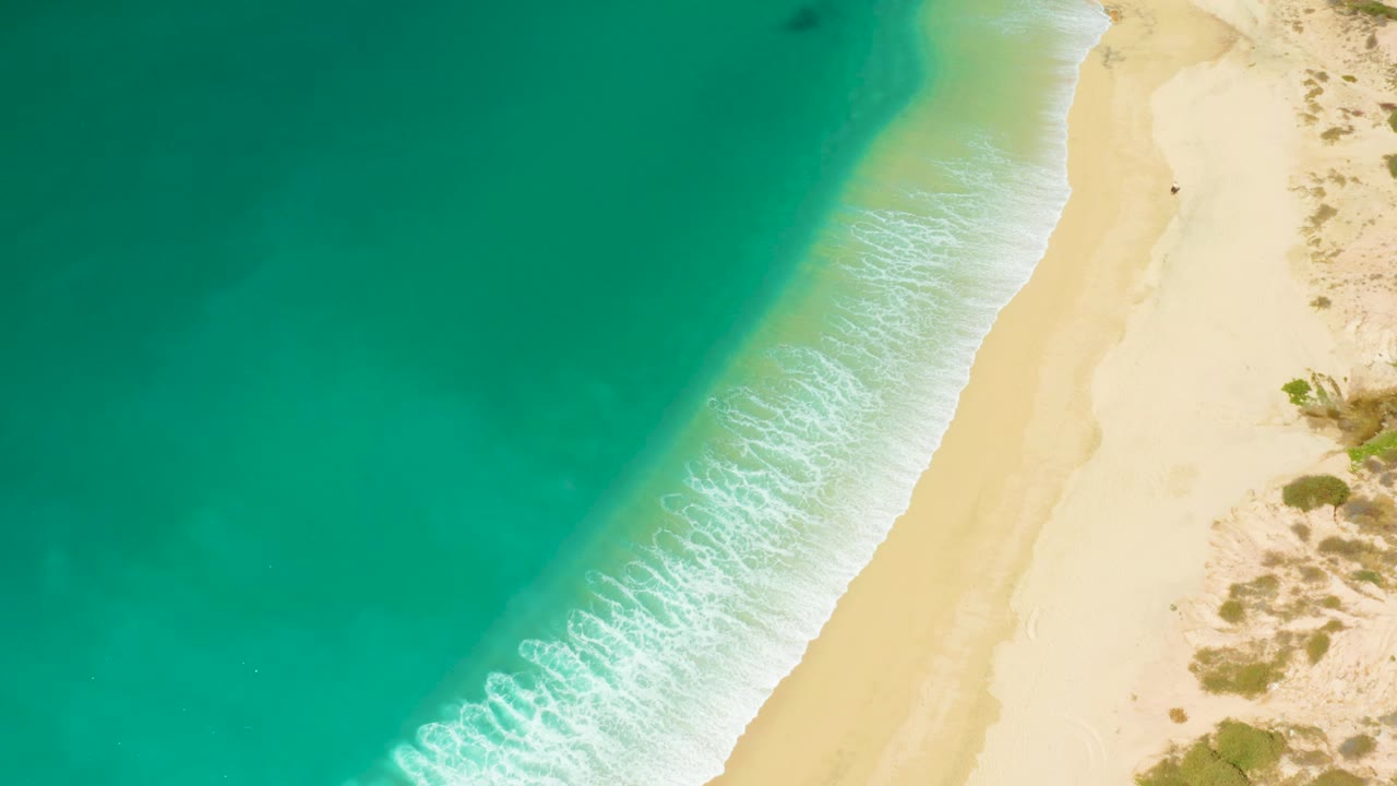 Top-down Hilton Los Cabos beach frame where the surf line sits lower in the view, turquoise water fills most of the left side, pale sand borders the right, and no visible people appear.