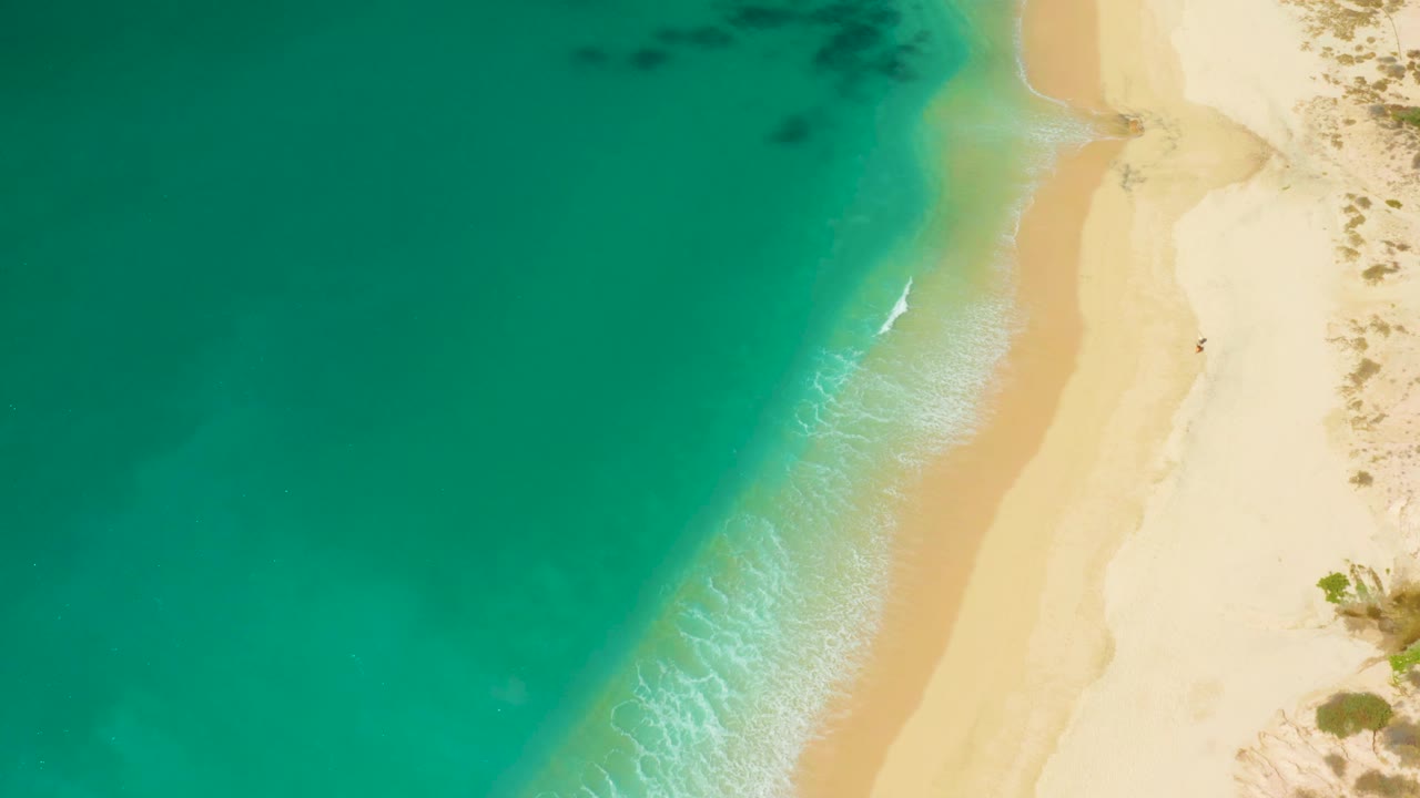 Aerial frame from Hilton Los Cabos, Mexico showing a slightly stronger white foam line where turquoise water meets the narrow sandy shore, with no visible people.