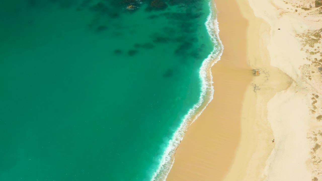 Aerial frame from Hilton Los Cabos, Mexico with the shoreline slightly shifted, dark underwater patches visible in the turquoise water, and no visible people.