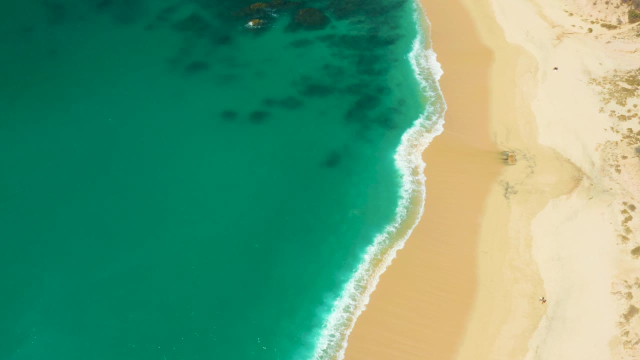 Overhead Hilton Los Cabos, Mexico shoreline view with green-blue water, pale sand, a curving band of wave foam, and no visible people.