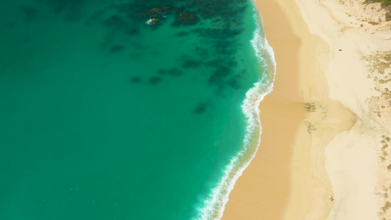 Drone-style overhead frame at Hilton Los Cabos, Mexico showing more of the water side, a vertical sandy shore, white wave wash, and no visible people.