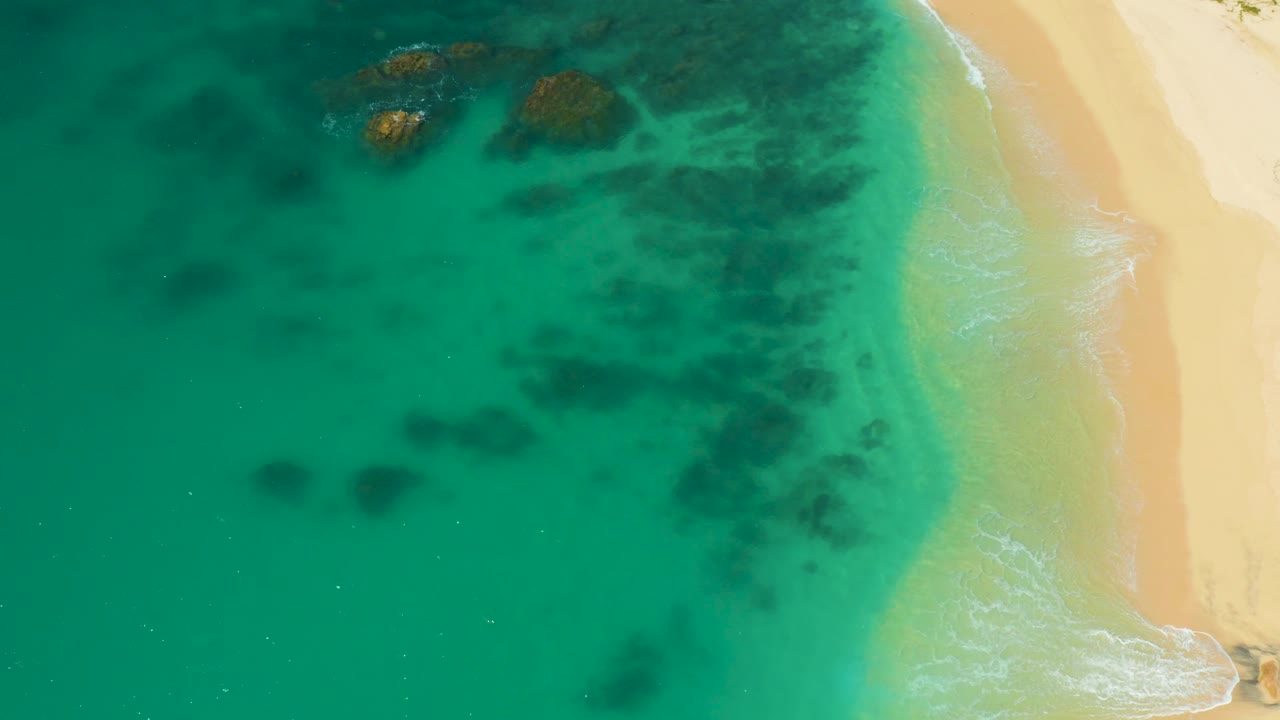 Aerial view over Hilton Los Cabos shallows with rocks near the top of frame, darker underwater areas across the water, pale beach on the right, and no visible people.