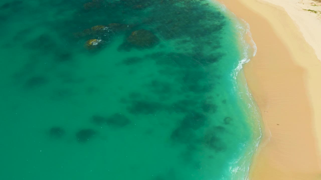 Aerial frame from Hilton Los Cabos, Mexico showing a wider sweep of turquoise ocean, pale sand at right, scattered darker underwater rock shapes, and no visible people.