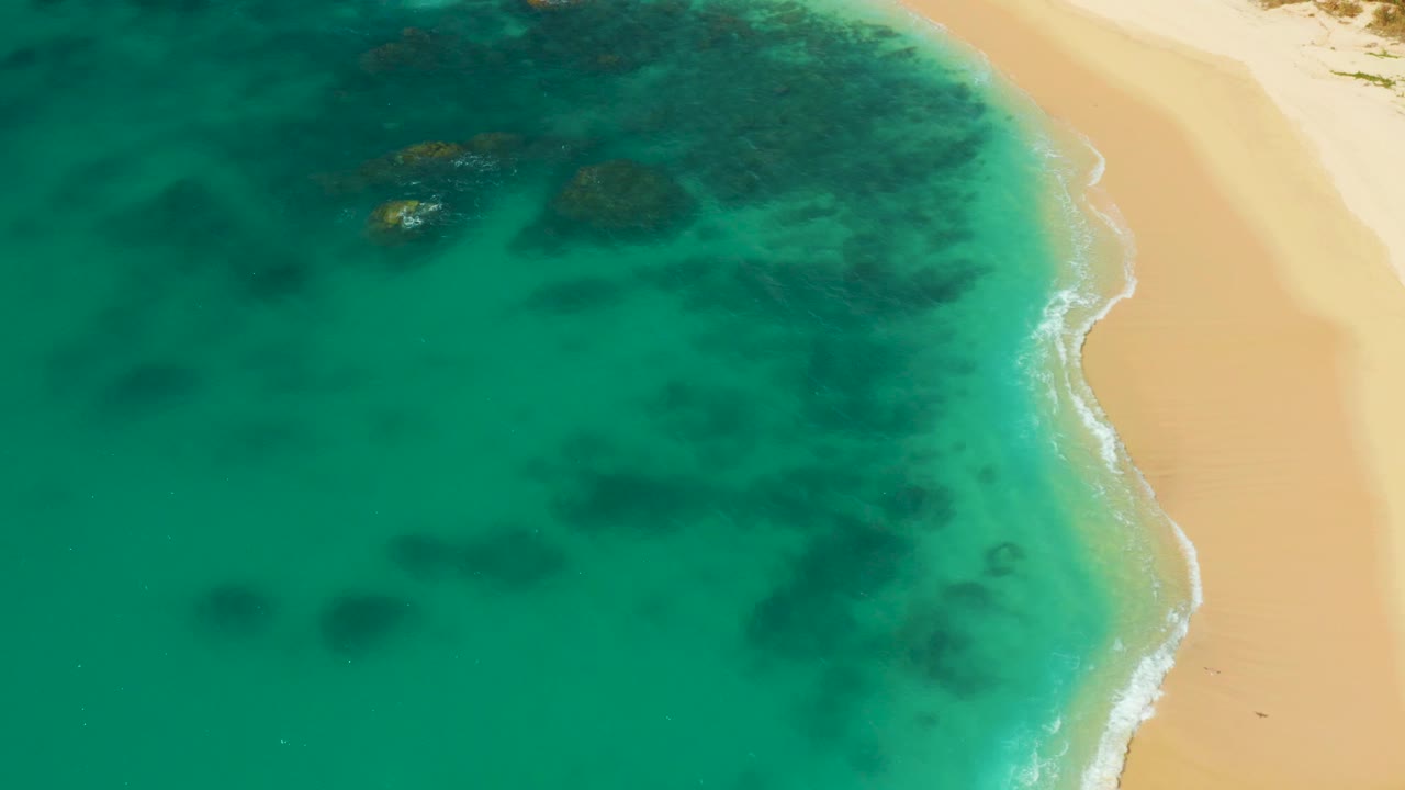 Overhead view at Hilton Los Cabos, Mexico with clear teal water on the left, sandy beach on the right, a narrow white surf line, and no visible people.