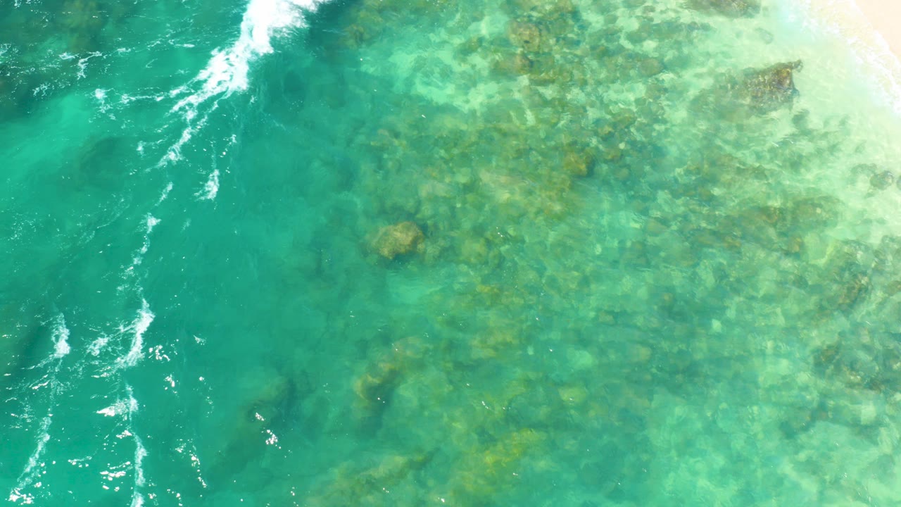 Overhead view at Hilton Los Cabos of clear turquoise shallow water showing a rocky seabed, with white surf foam entering along the upper left edge and no visible people.