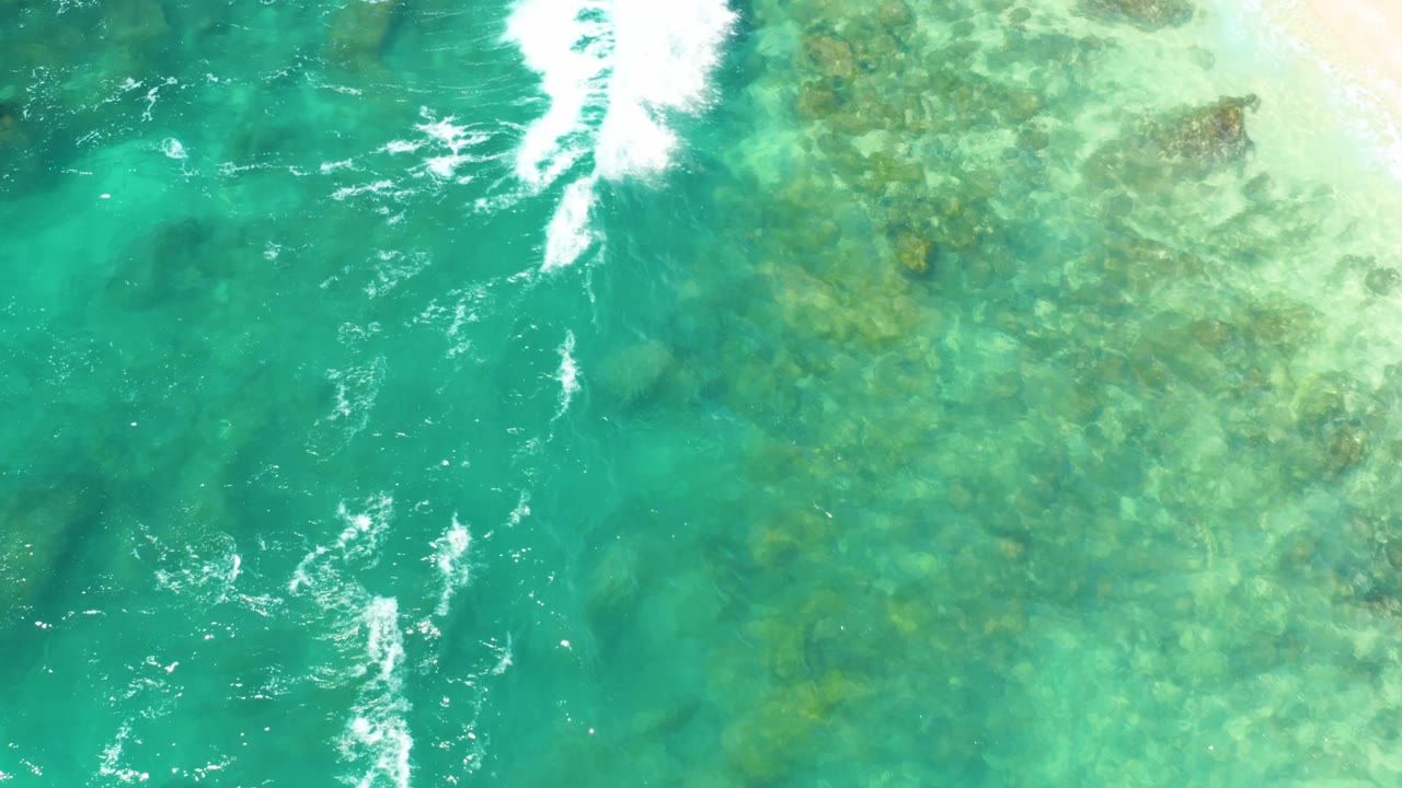 Overhead view at Hilton Los Cabos with bright turquoise water, mottled rocky seabed below the surface, foamy white surf near the top edge, and no visible people.