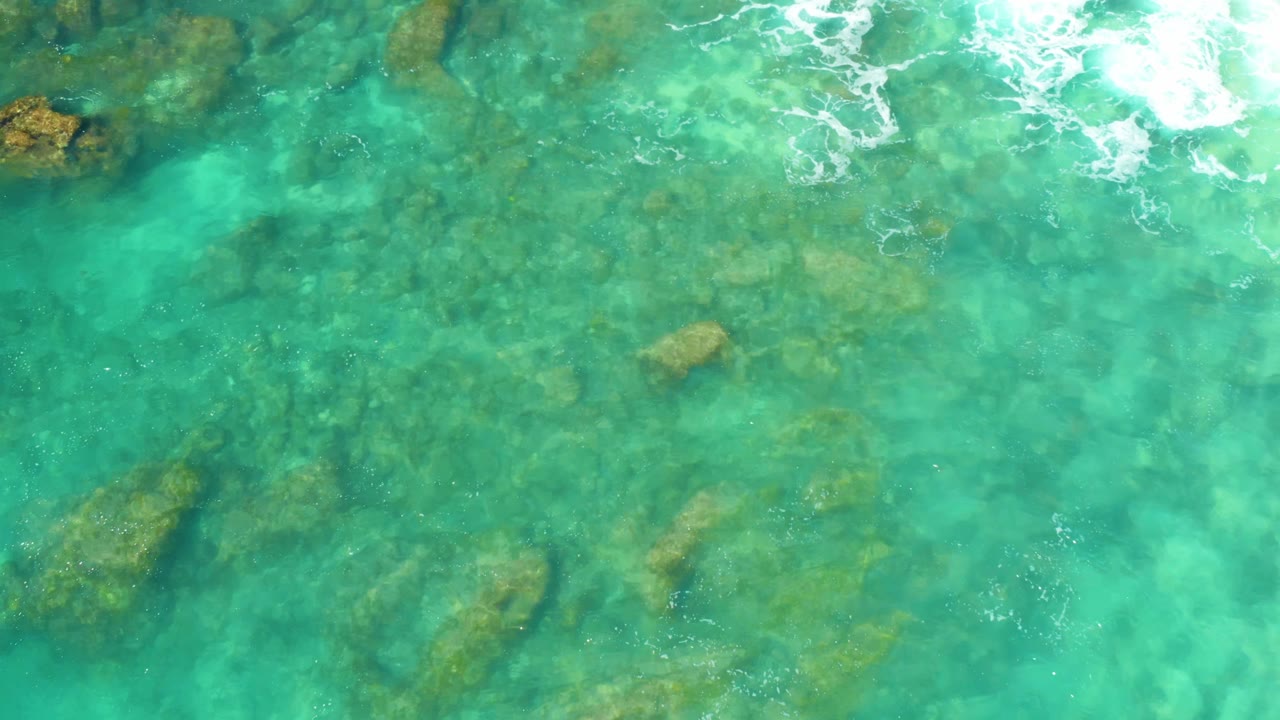 Clear green-blue seawater at Hilton Los Cabos, Mexico, shows scattered submerged rocks beneath small ripples, with foam shifted along the upper frame and no visible people.