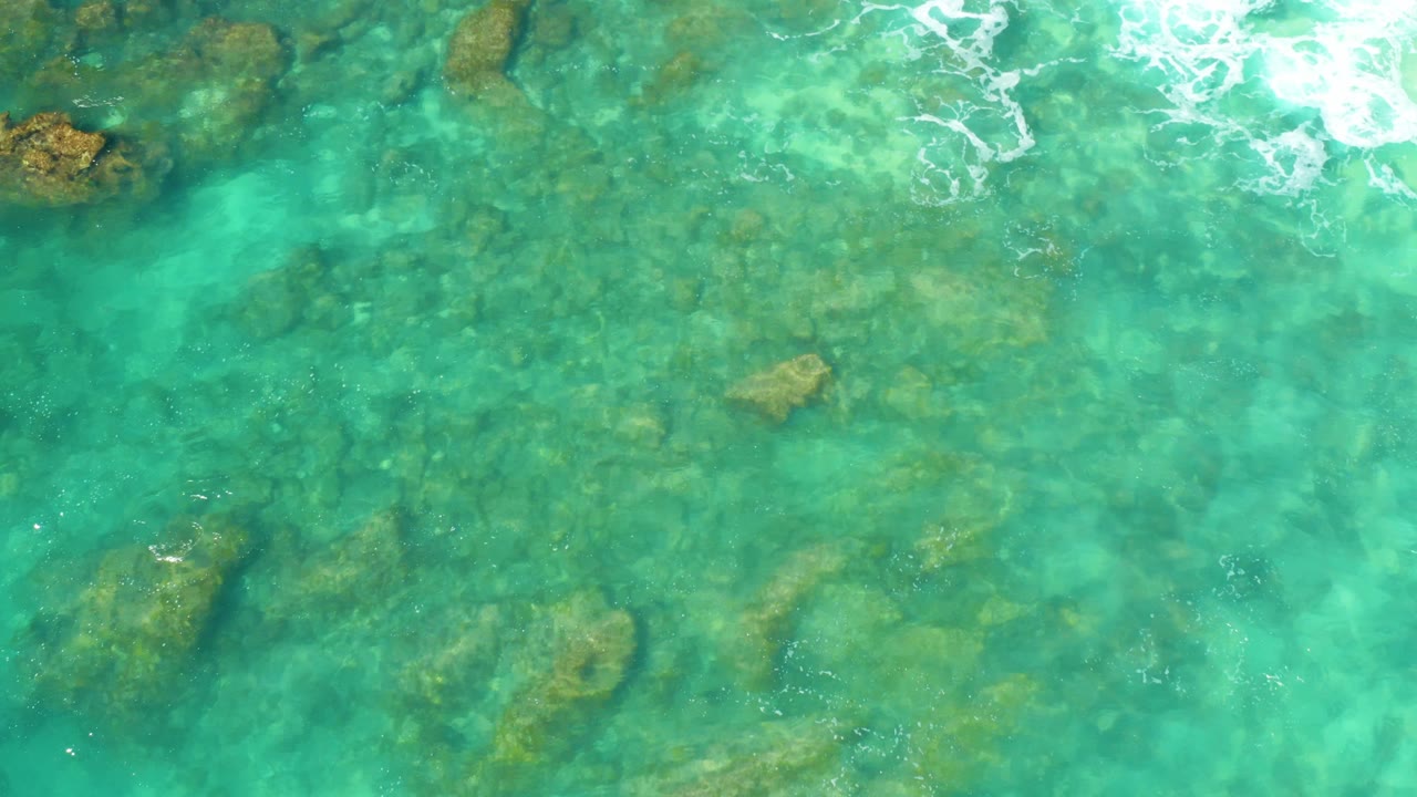A top-down frame at Hilton Los Cabos, Mexico, shows shallow turquoise water, scattered underwater rocks, and a more distinct rock near the upper left, with no visible people.
