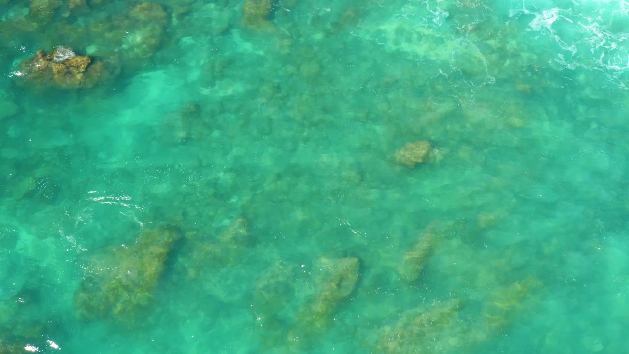 Clear turquoise water at Hilton Los Cabos fills the frame, with darker underwater rock shapes and small surface ripples, no visible people.