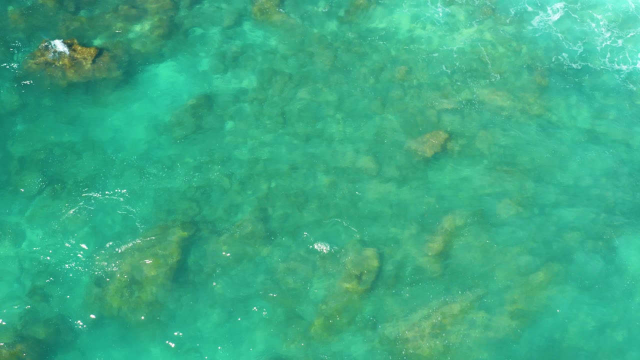 Overhead frame from Hilton Los Cabos with bright turquoise shallows, muted submerged rocks, and a small patch of white water texture near the upper edge, no visible people.
