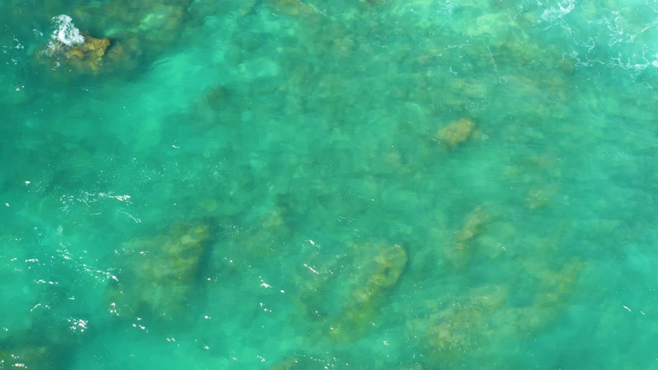 Top-down Hilton Los Cabos water view with a larger submerged rock area crossing the center and light ripples on the turquoise surface, no visible people.