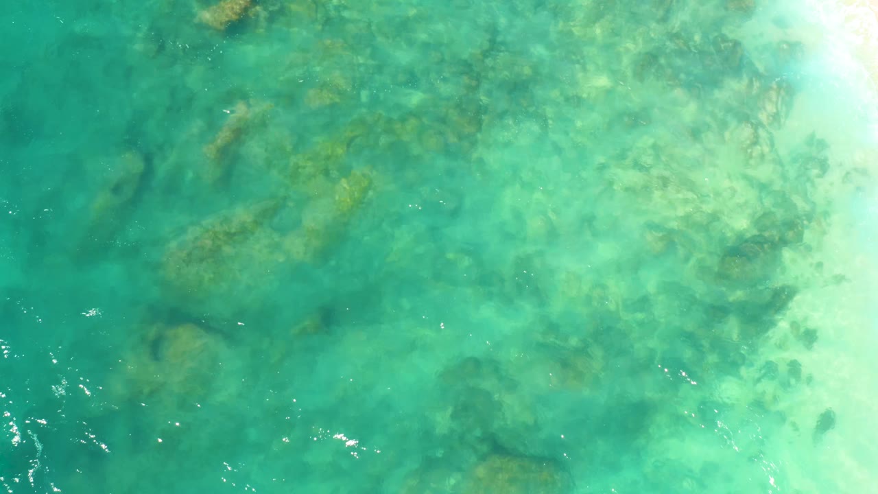 Aerial-looking view of green-blue ocean water at Hilton Los Cabos, Mexico, with darker submerged rock patches beneath rippled surface reflections, no visible people.