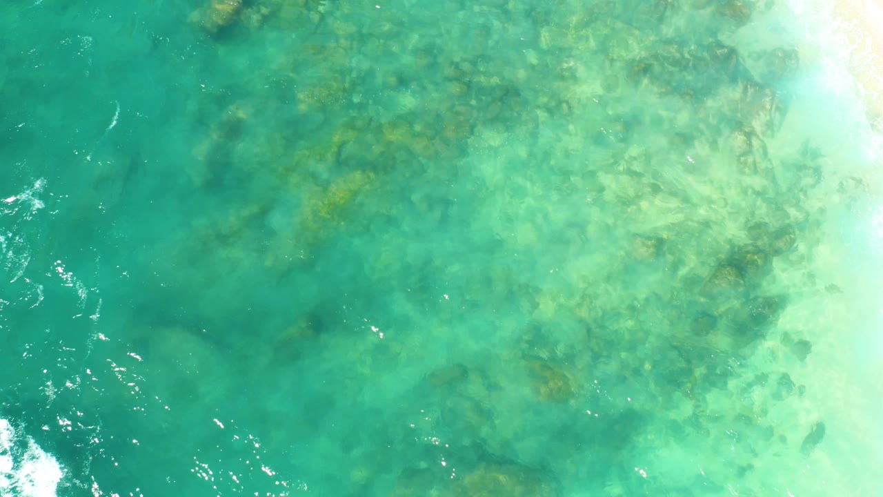 Clear turquoise water at Hilton Los Cabos, Mexico, reveals uneven submerged rocks while bright white foam and pale water sit along the right side, no visible people.