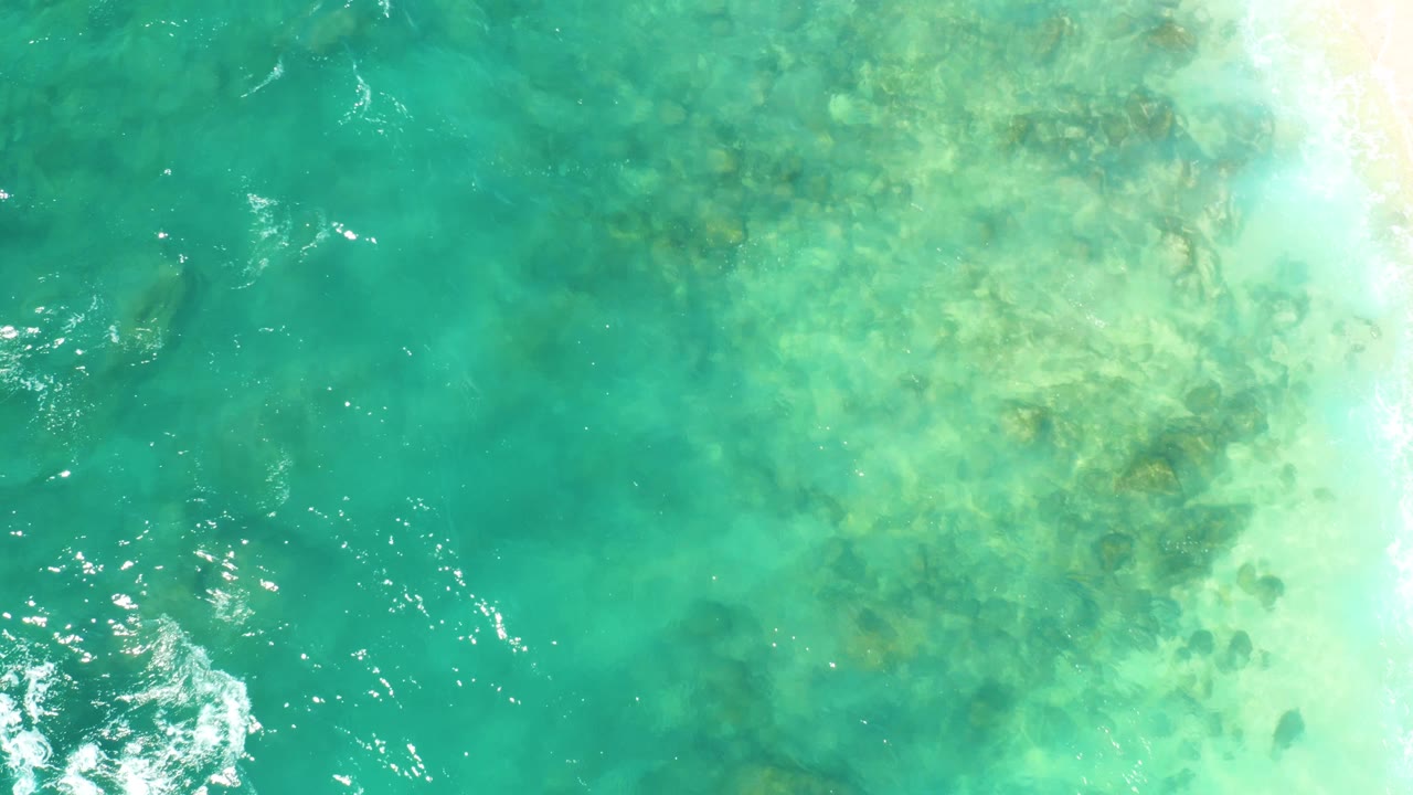 Top-down frame from Hilton Los Cabos, Mexico, showing shallow turquoise water, scattered submerged rocks, and small bright reflections near the left and lower edges, no visible people.