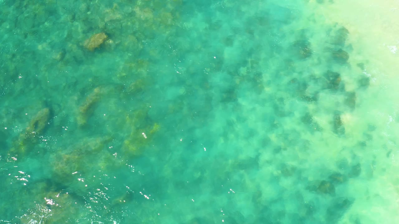 Top-down frame from Hilton Los Cabos showing blue-green shallow water, scattered underwater rocks, small bright reflections, and no visible people.