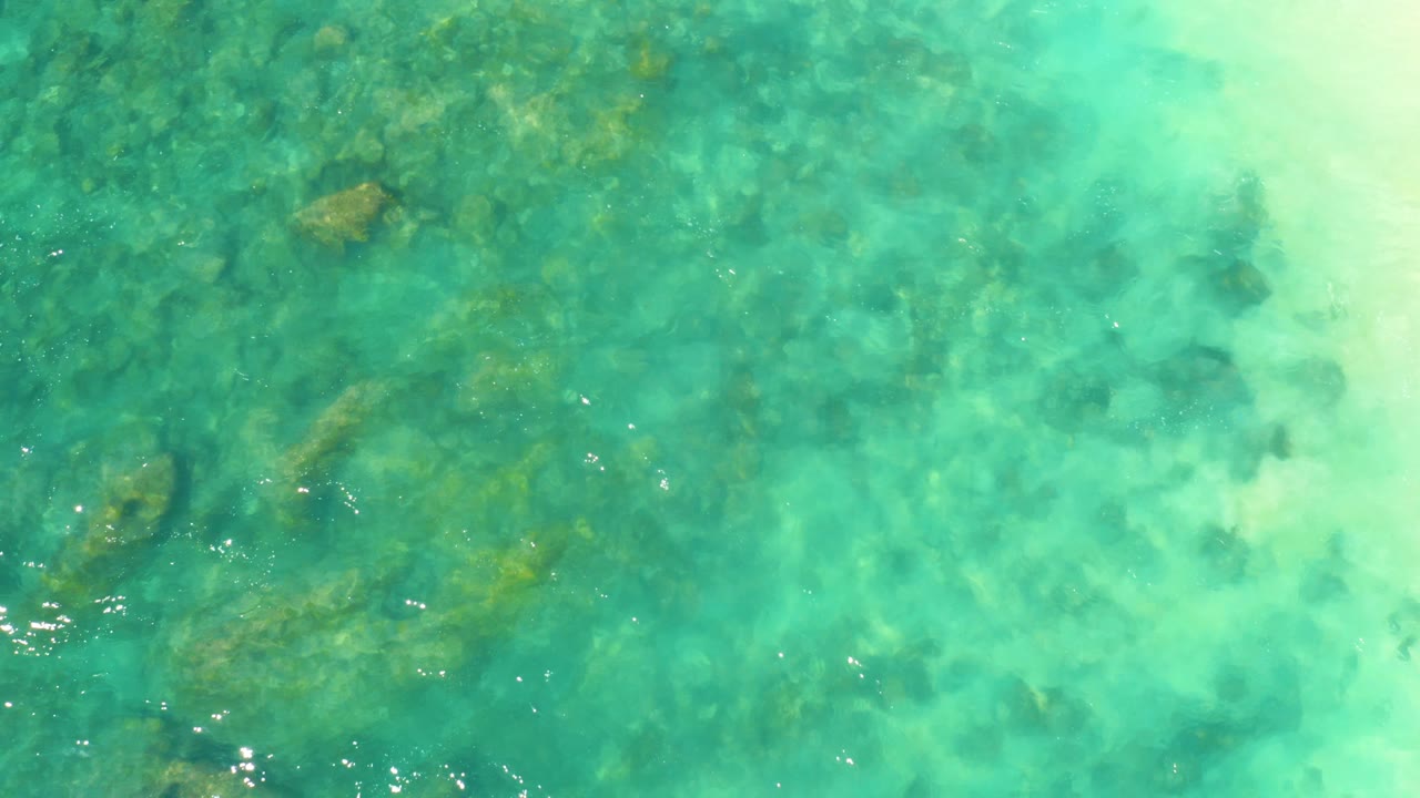 Overhead Hilton Los Cabos water detail with a denser rocky seabed visible through green-blue shallows and no visible people.