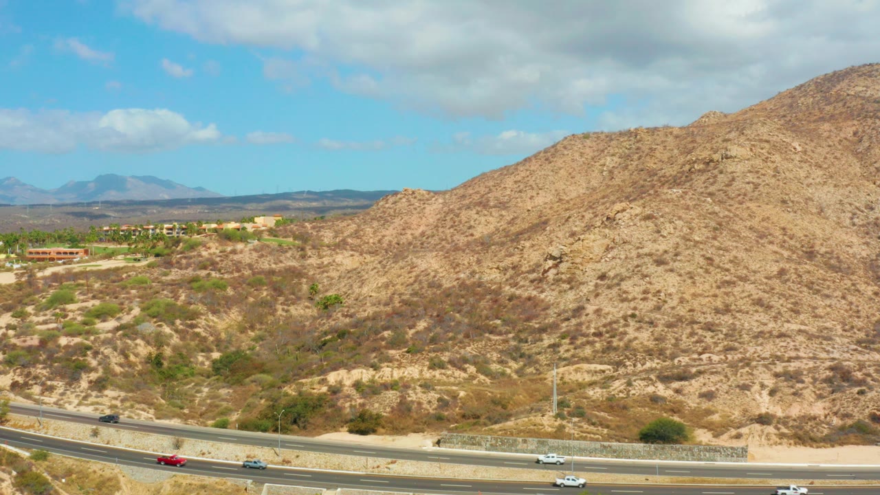 At 0:00, an elevated view near Hilton Los Cabos, Mexico shows cars on a multi-lane highway below tan dry hills, distant buildings, blue sky, and no visible people.