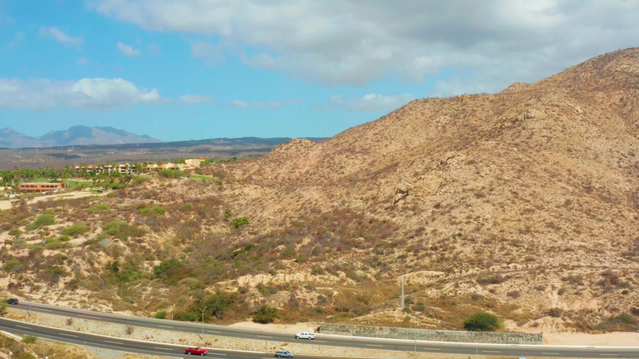 At 0:02, a red car stands out on the highway foreground, with tan hills, scattered green shrubs, distant structures, and partly cloudy sky near Hilton Los Cabos, Mexico, with no visible people.
