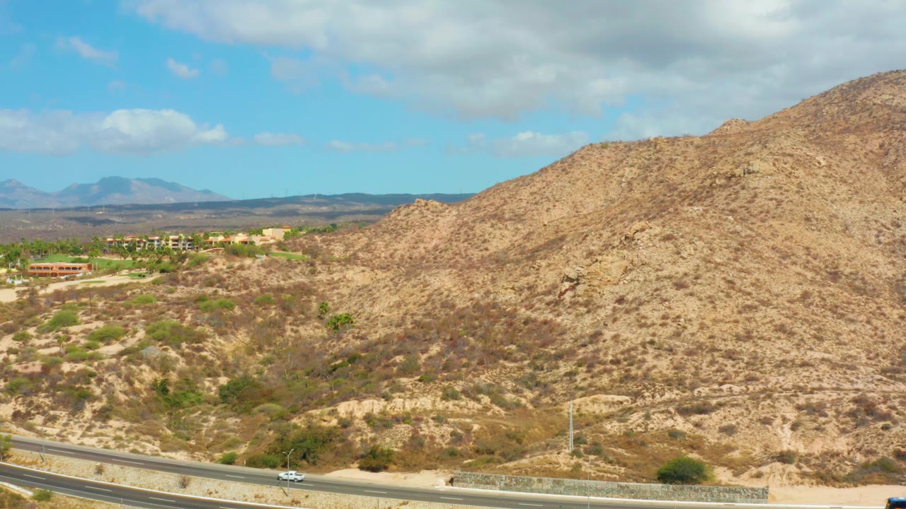 At 0:03, the highway runs across the bottom of the frame while the hillside fills more of the view, with small buildings to the left and no visible people near Hilton Los Cabos, Mexico.