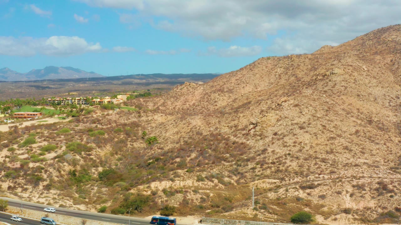 Opening frame of Hilton Los Cabos, Mexico desert terrain with a highway at the bottom, tan scrub hills, distant mountains, blue sky, and no visible people.