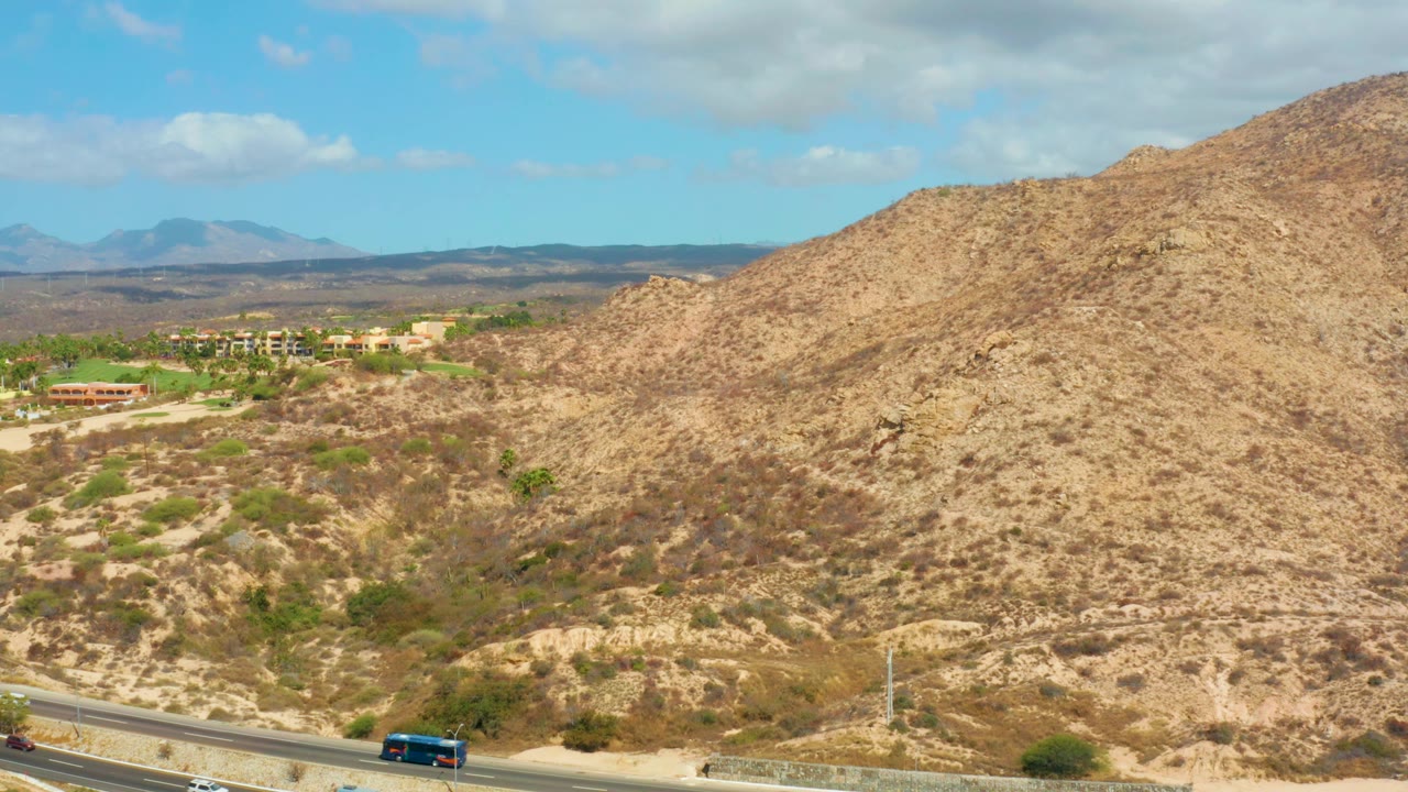 Third frame catches a blue vehicle lower right on the Hilton Los Cabos, Mexico road, with dry hills, scattered shrubs, distant ridges, and no visible people.
