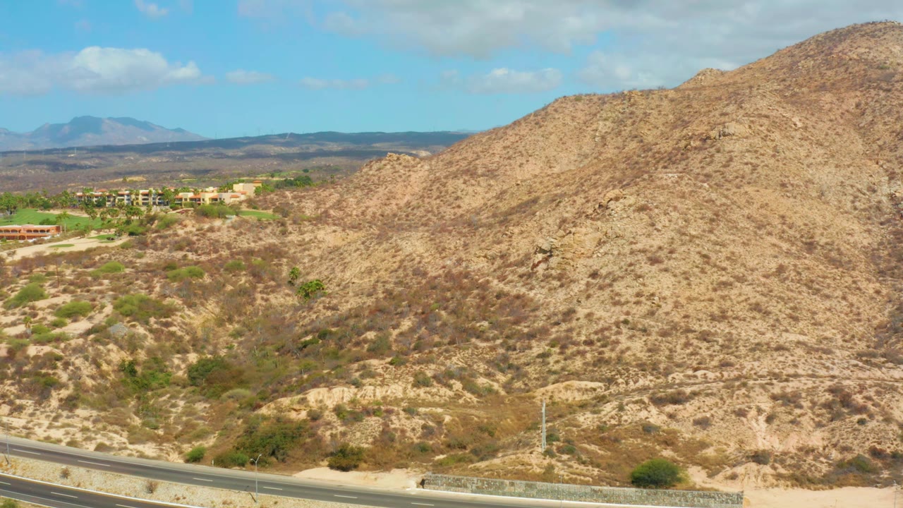 Elevated Hilton Los Cabos landscape frame with the highway still visible along the lower left, tan scrub-covered hills dominating the view, distant buildings, and no visible people.