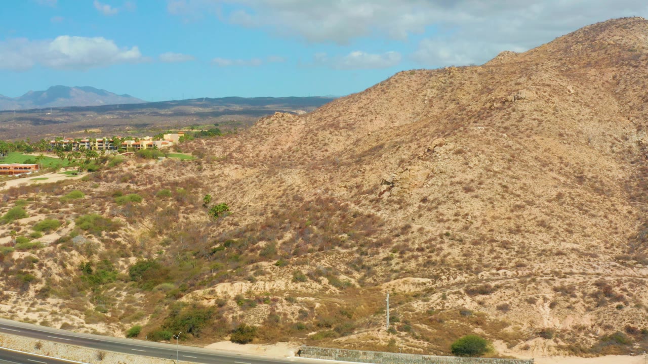 Aerial view near Hilton Los Cabos with the road compressed along the bottom edge, a broad tan hill across the center and right, distant mountain shapes, and no visible people.