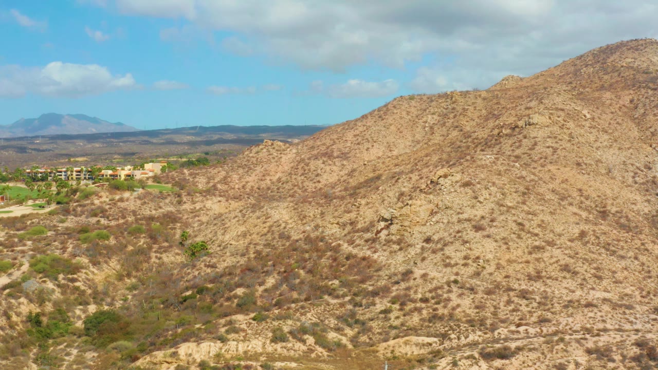 A high view over a dry hillside near Hilton Los Cabos, Mexico, shows scrubby tan slopes, a small cluster of distant buildings on the left, hazy mountains, and no visible people.