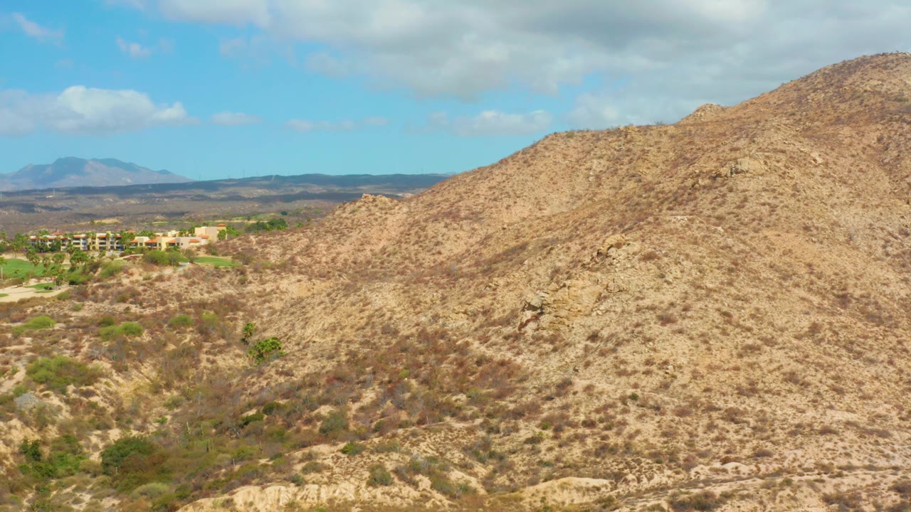 The hillside remains centered near Hilton Los Cabos, Mexico, with rocky tan ground, scattered shrubs, distant structures at the left edge, soft clouds above, and no visible people.