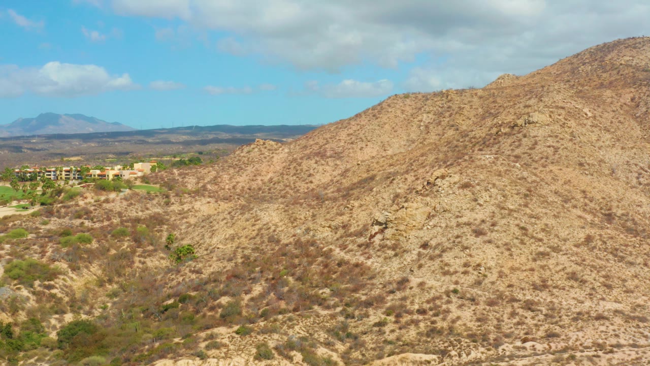 Mid-clip view of the arid hillside near Hilton Los Cabos, Mexico, with sparse green patches in the dry wash, distant buildings to the left, mountains on the horizon, and no visible people.