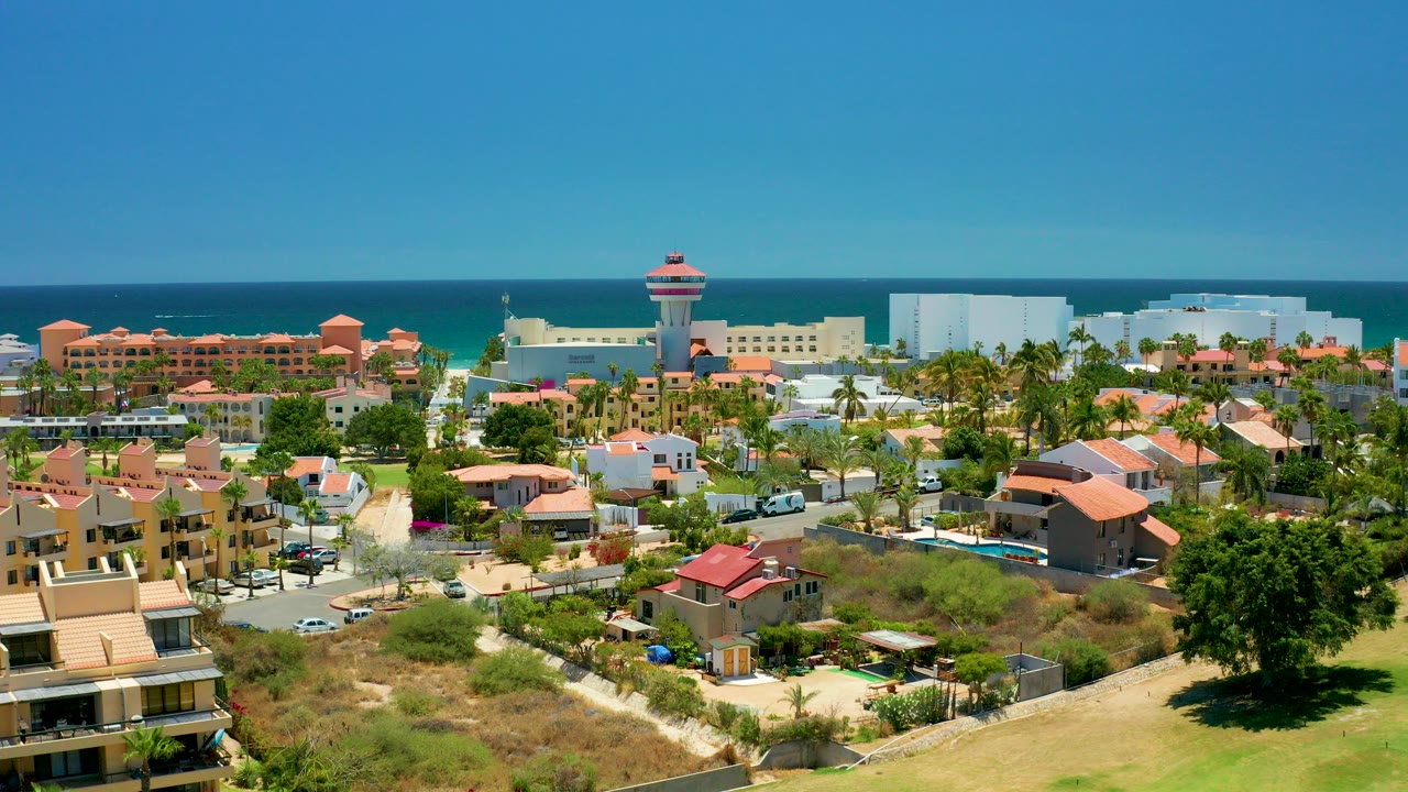 Wide aerial view over San Jose Del Cabo homes with red tile roofs, palm trees, parked and moving cars, a rounded tower-like building near the shore, ocean beyond, and no visible people.