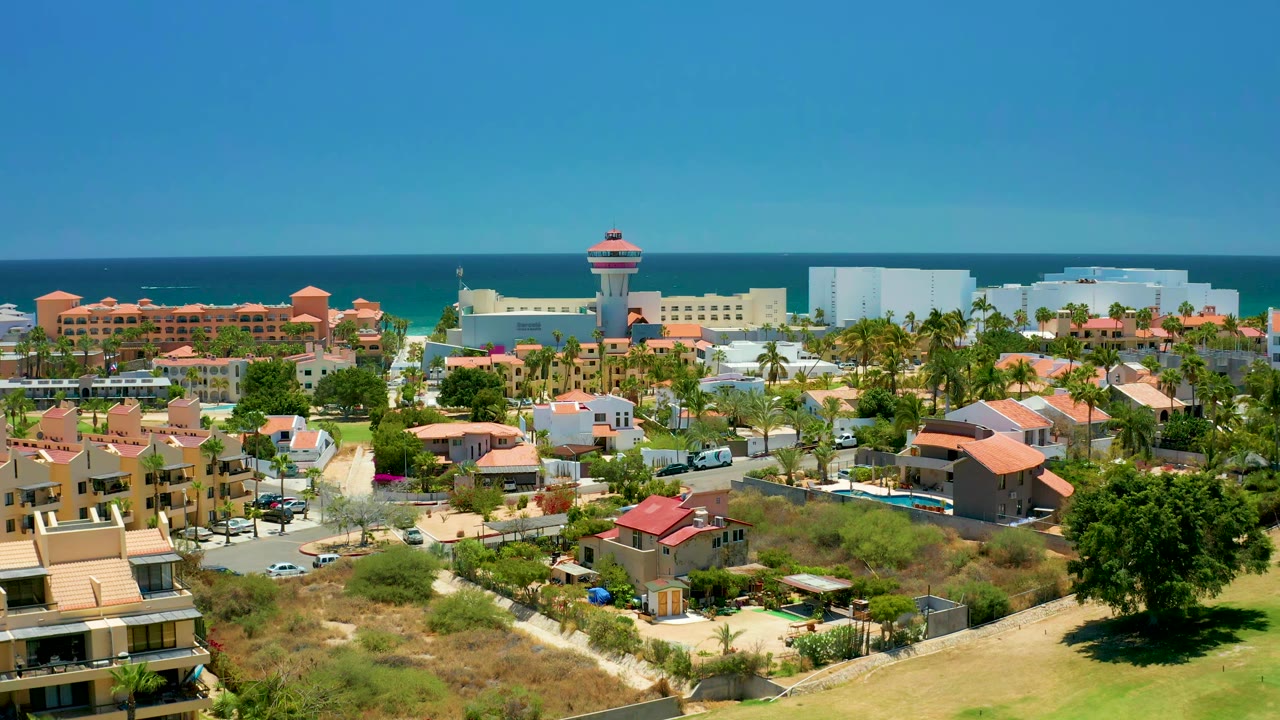 Coastal neighborhood with red-roofed buildings, roads, cars, palms, and ocean horizon.