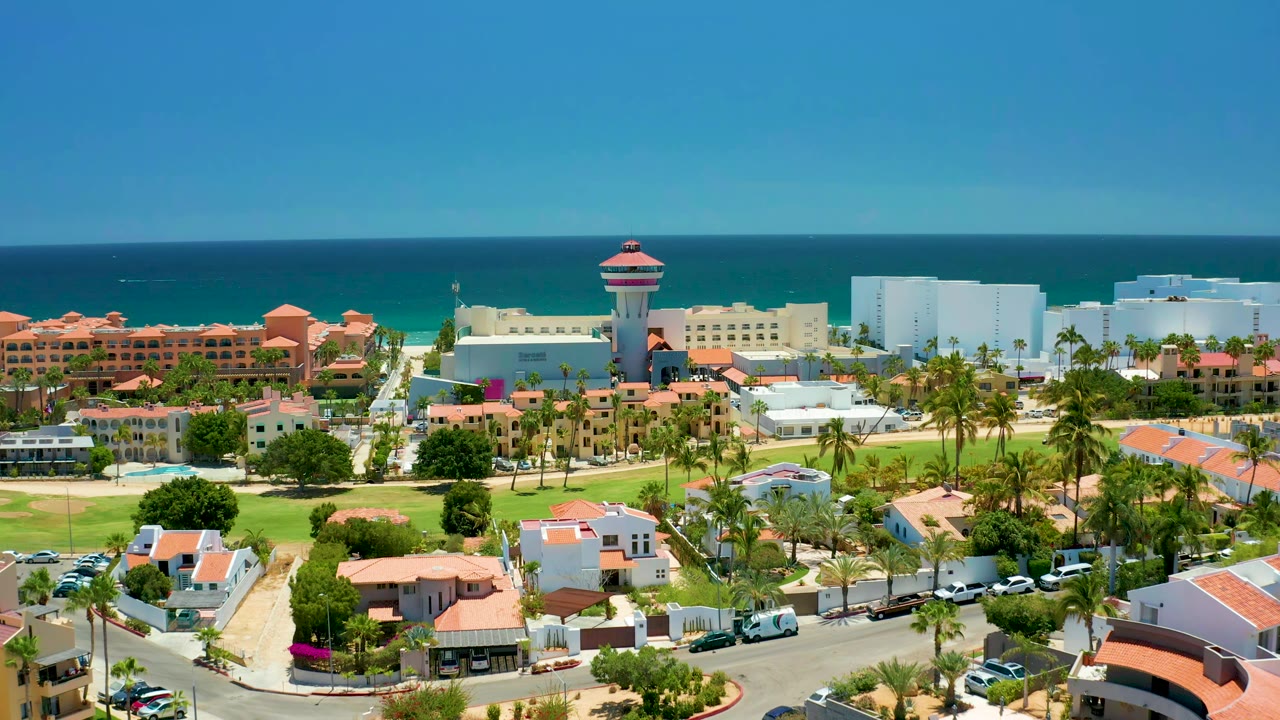 Aerial view across resort rooftops, palms, hotel blocks, and blue ocean.