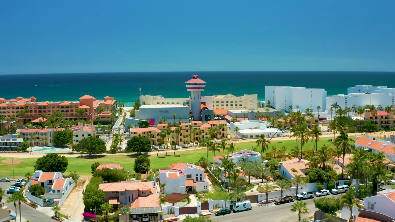 Aerial frame over San Jose Del Cabo coastal development with resort roofs, a curved foreground road, parked and moving cars, palm trees, ocean water, and no visible people.