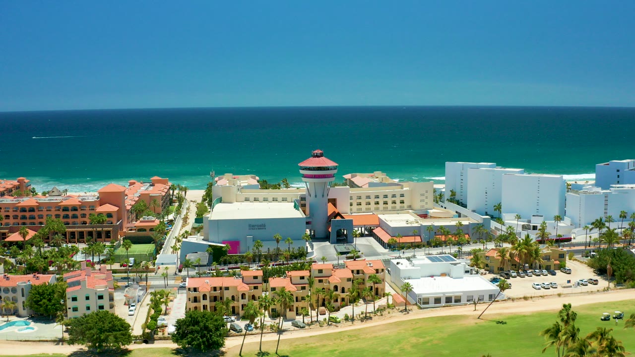 Opening aerial frame over San Jose Del Cabo coastal buildings, palm trees, a round water tower, turquoise ocean, and clear blue sky with no visible people.