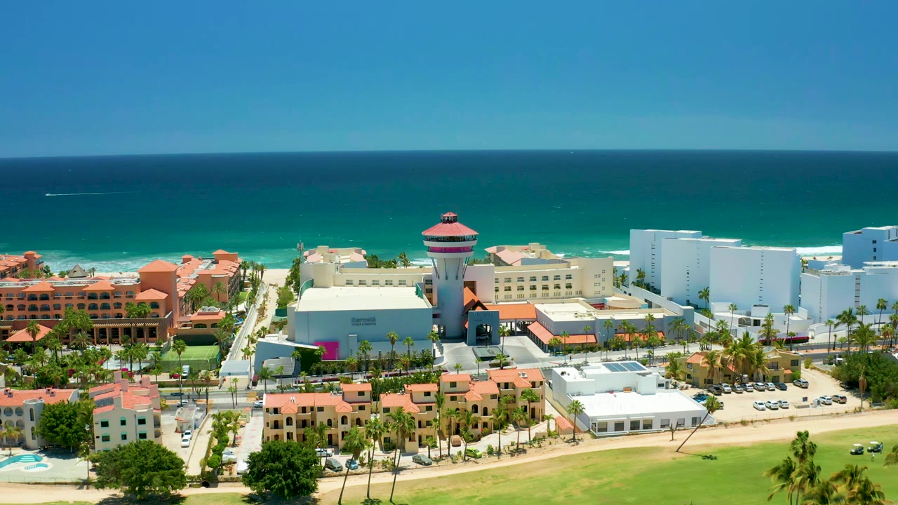Aerial view one second into the clip showing San Jose Del Cabo beachfront development, terracotta roofs, white buildings, palm rows, and ocean surf with no visible people.