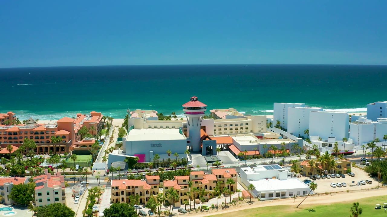 San Jose Del Cabo aerial frame with the water tower near center, white coastal buildings to the right, orange-roofed structures to the left, and waves along the beach with no visible people.